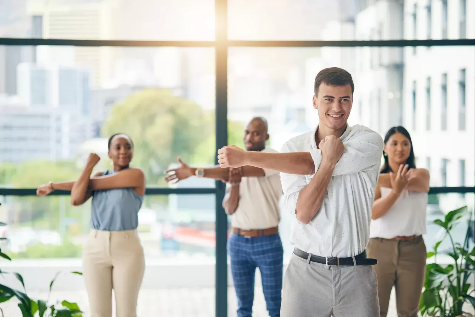 A group of diverse business professionals stretching together in an office setting, smiling and performing arm stretches in front of large windows with a city view